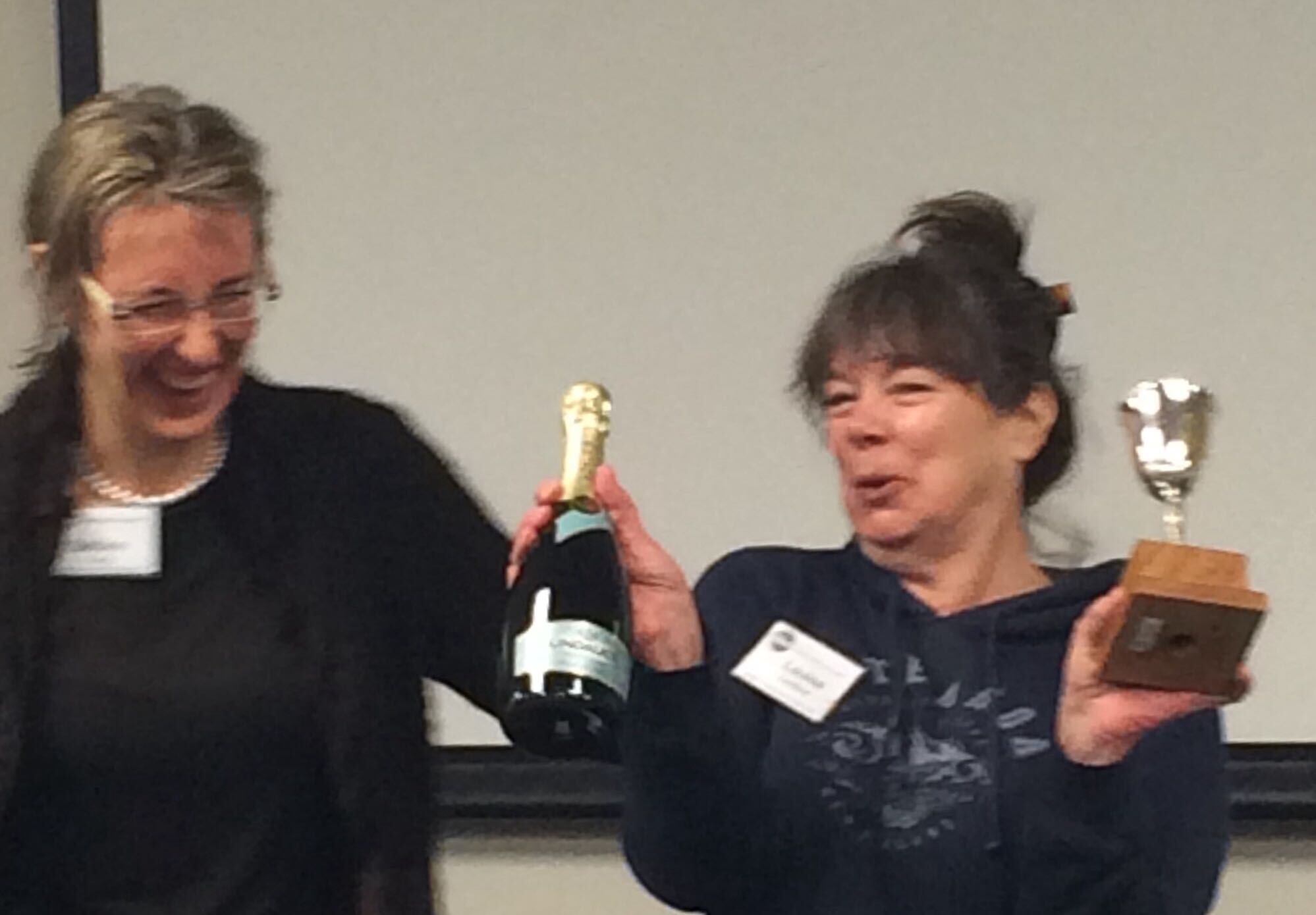 Two women laughing together, one holding a trophy and a bottle of wine