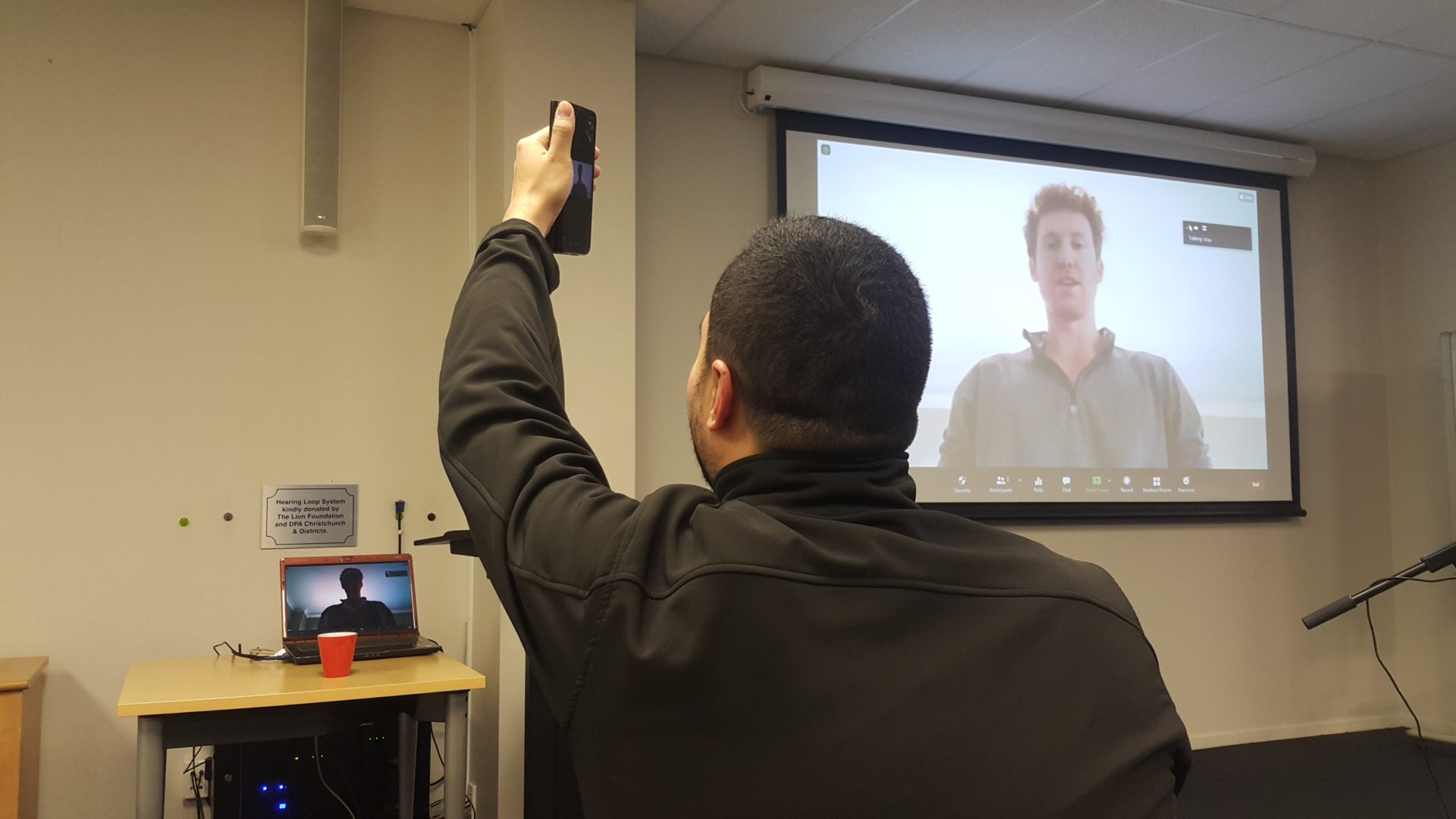 Man taking photo of person presenting on a large screen via Zoom