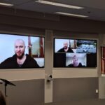 Two large digital displays. One showing a close up of the speaker Zooming in, the other showing him, plus a shot of the meeting room and the other online participant