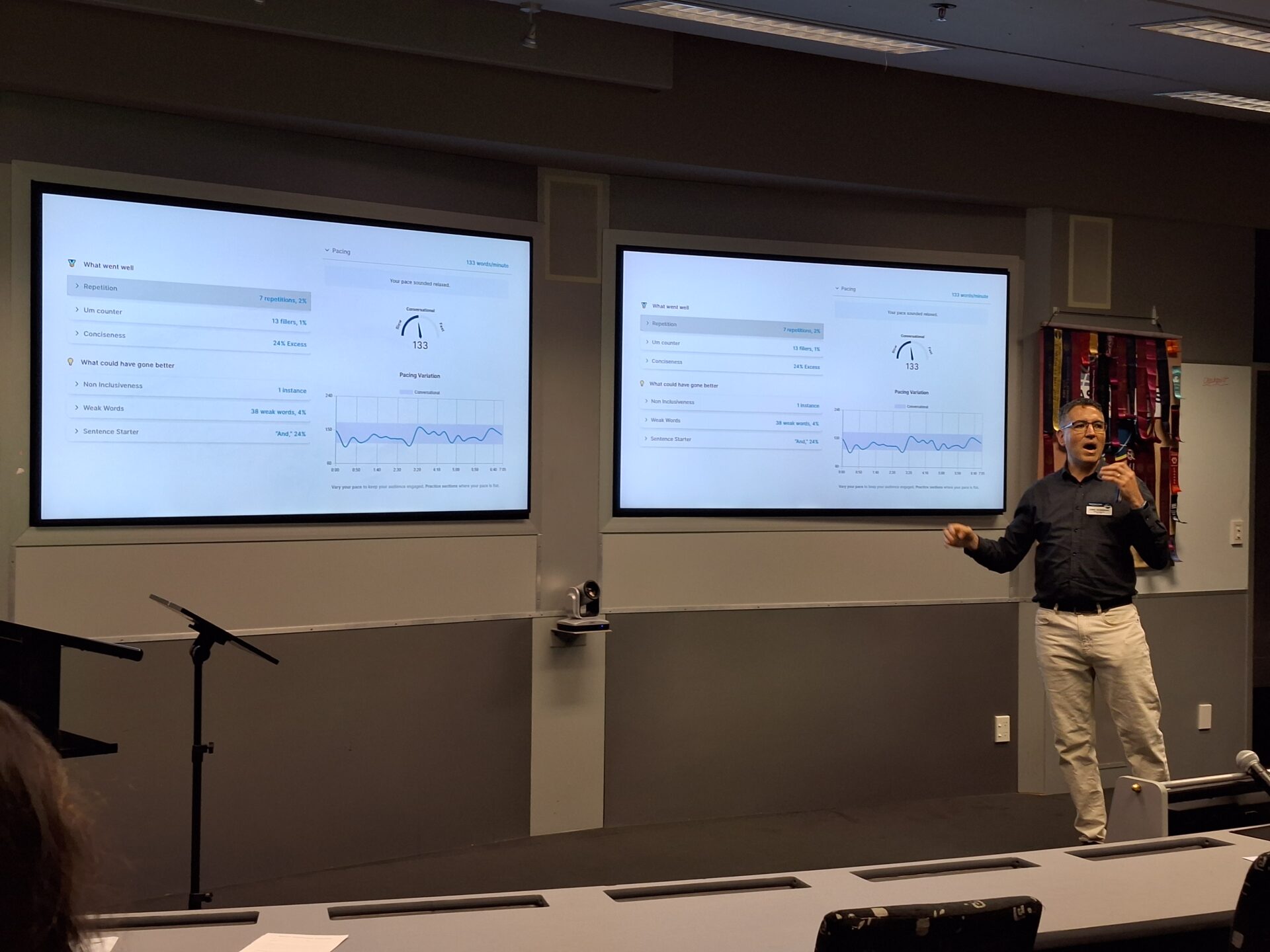 Man delivering a presentation in front of two large monitors displaying a Yoodli digital dashboard