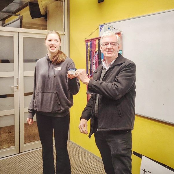 A young woman and an older man stand side by side in a room with a yellow wall and a whiteboard. The woman is wearing a grey hoodie and smiling, while the man is dressed in a dark jacket and glasses, also smiling. They are holding a small item together, possibly a card or plaque.