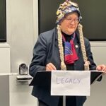 Woman wearing a bucket hat covered in badges and with two false long blonde plaits attached, standing behind a lectern