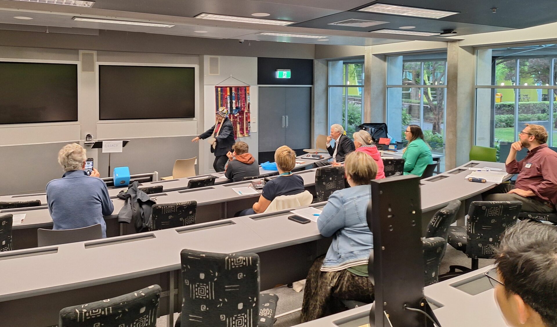 View from the back of a tiered lecture theatre showing seven or eight people from behind, looking at a woman wearing a wig with two blonde plaits, who is performing a speech