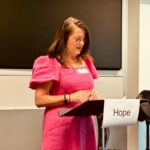 Woman wearing bright pink dress reading from a lectern, in front of which hangs a sign with the word Hope written on it