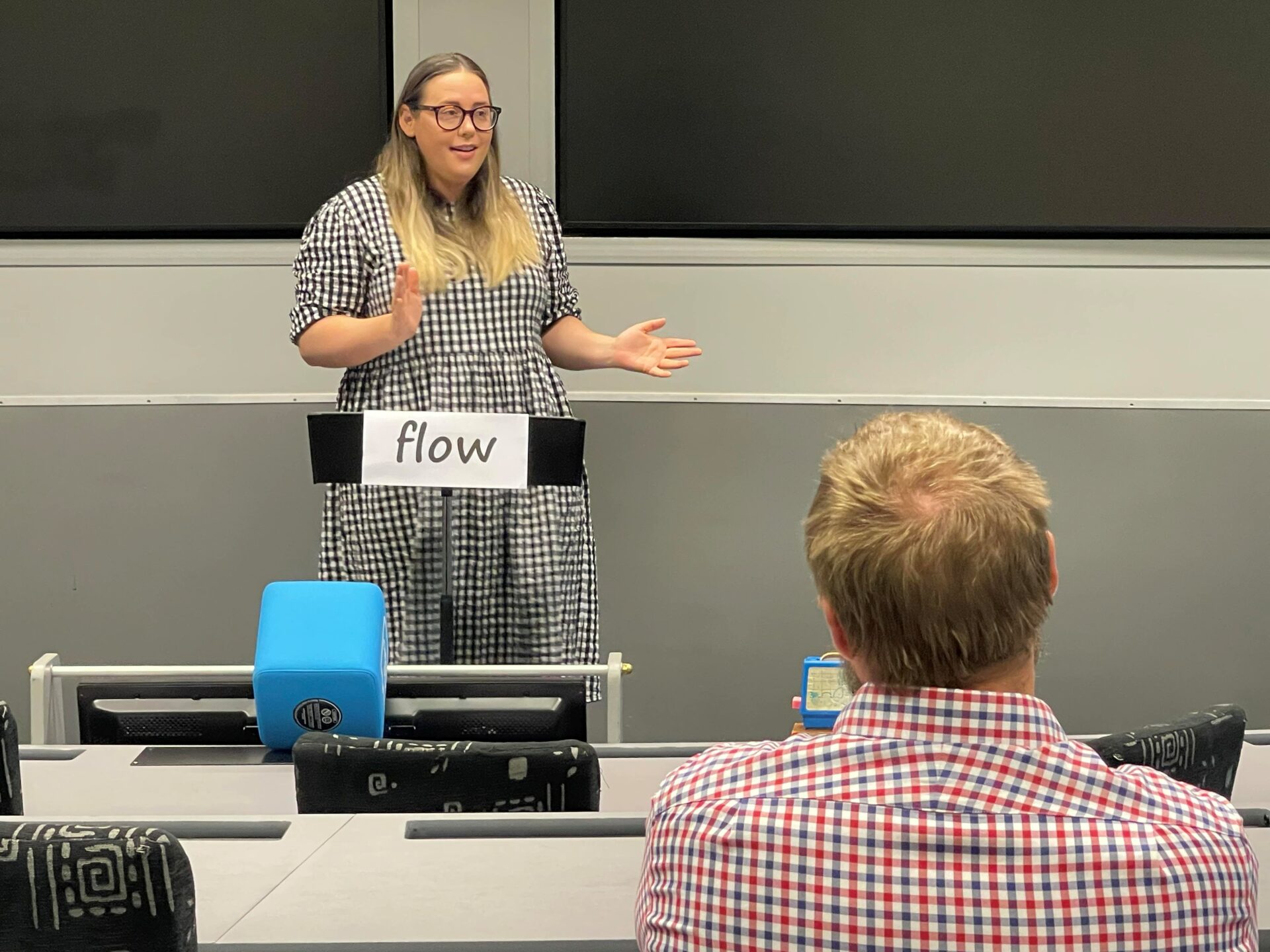 Woman in a black and white checkered dress standing behind a lectern which has the word flow written on a piece of paper, hanging from the front of it, facing an audience. She is giving a speech.