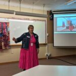 Woman in a bright pink dress and denim jacket stading in front of a whiteboard and powerpoint presentation giving a speech