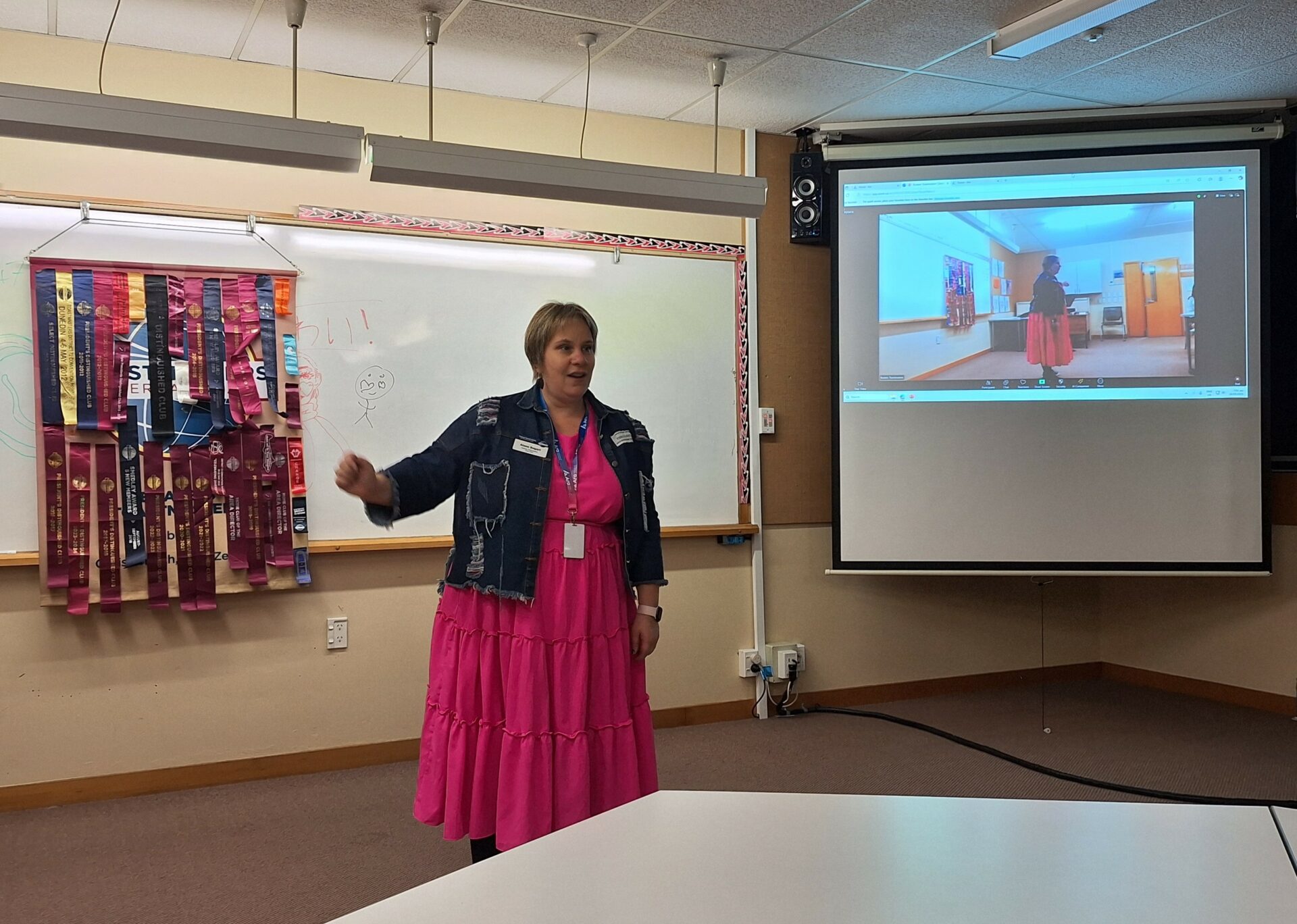 Woman in a bright pink dress and denim jacket stading in front of a whiteboard and powerpoint presentation giving a speech