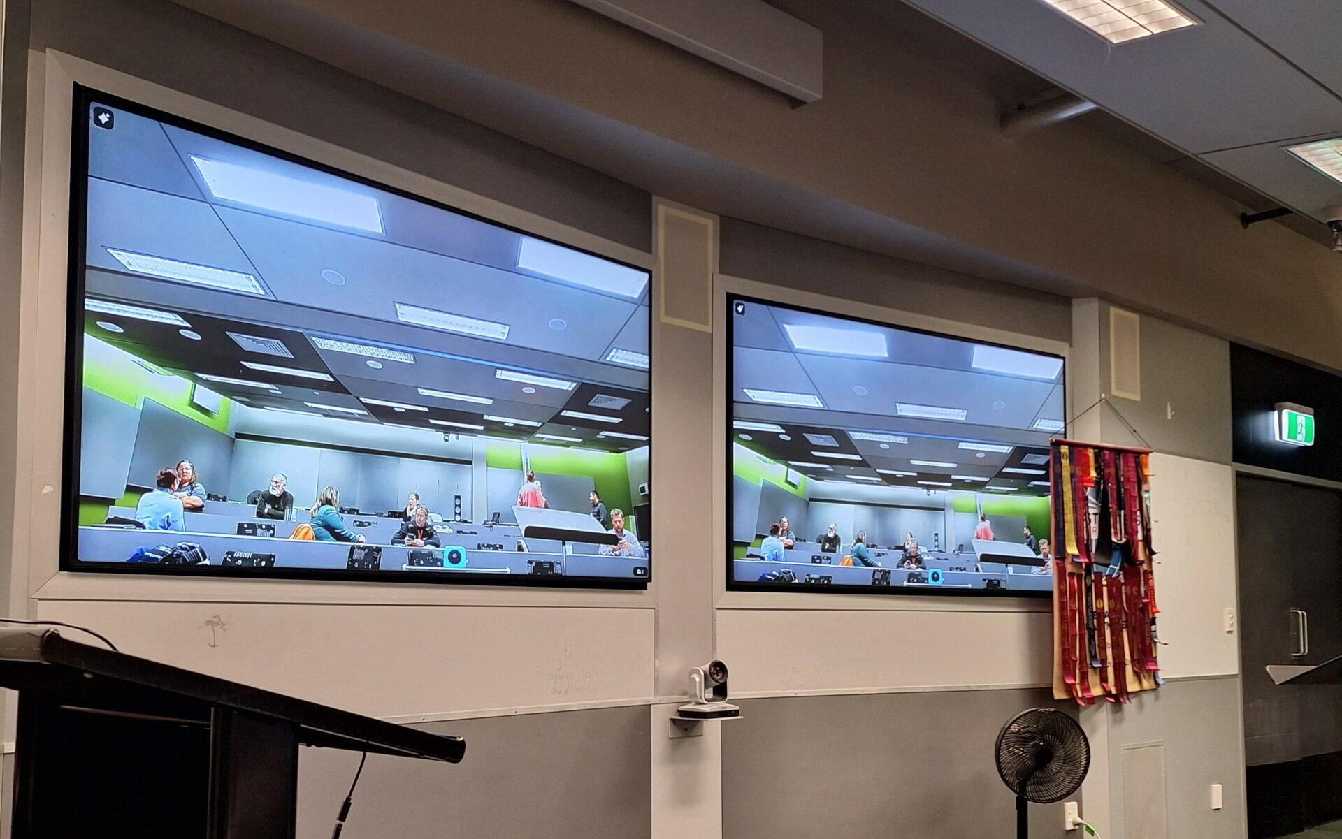 View of two large presentation screens showing lecture theatre with people chatting to each other around the rows